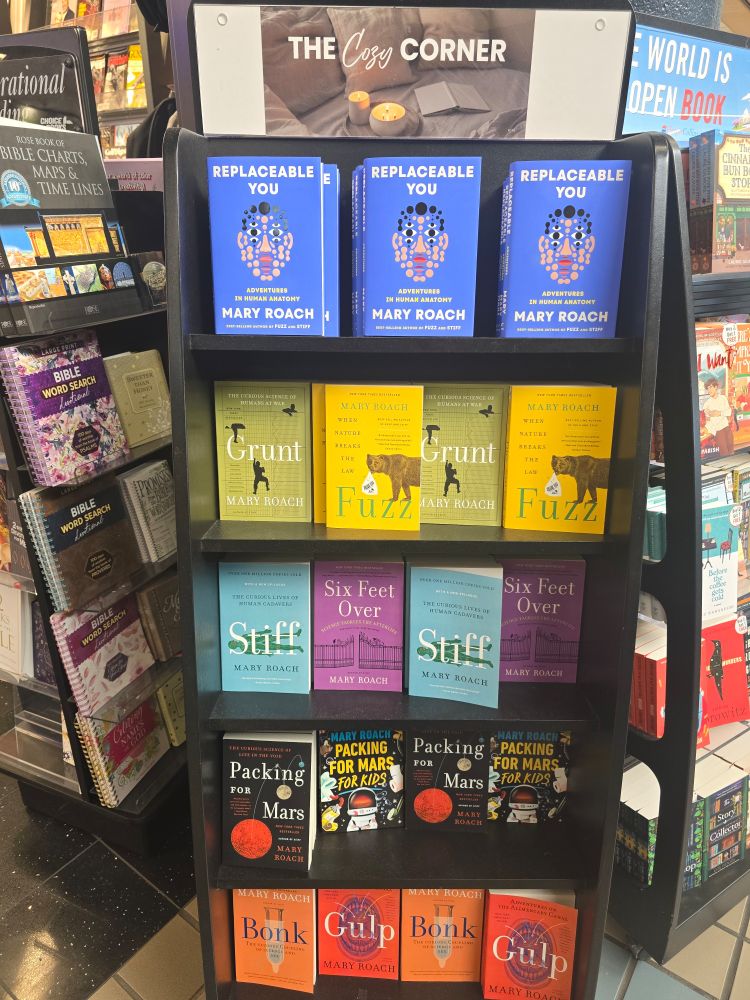 A bookshelf in an airport shop labeled "cozy corner" with a bunch Mary Roach's books