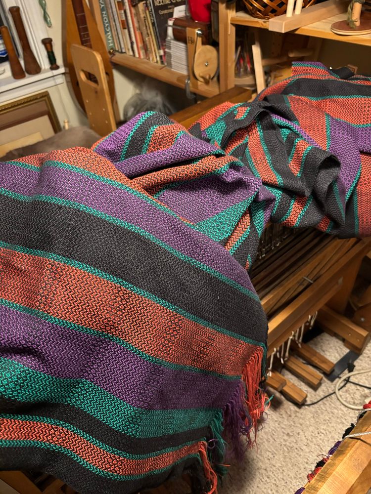 Handwoven yardage fresh from the loom in a chevron and rose path point twill. Colors are black, green, orange and purple. It is all piled on top of a floor loom with bookshelves behind. 