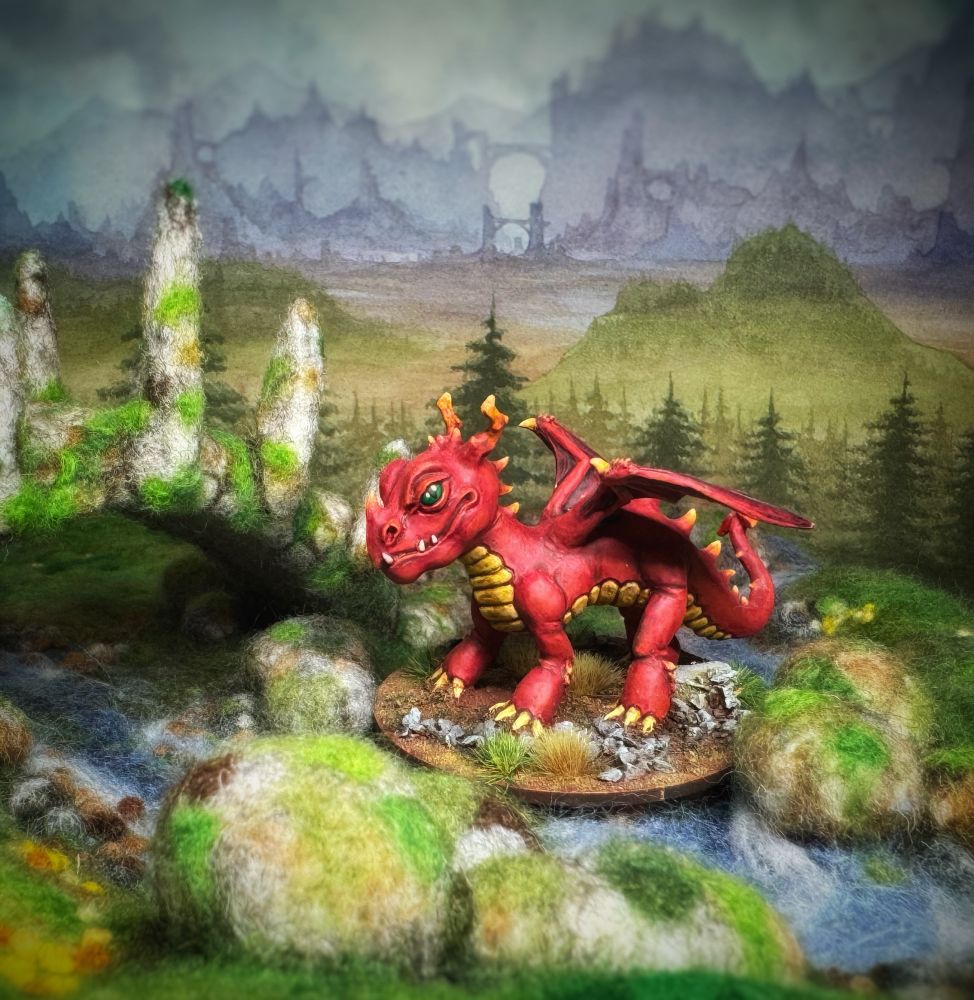 A painted red dragon miniature on needle felted terrain with a painted landscape background.

