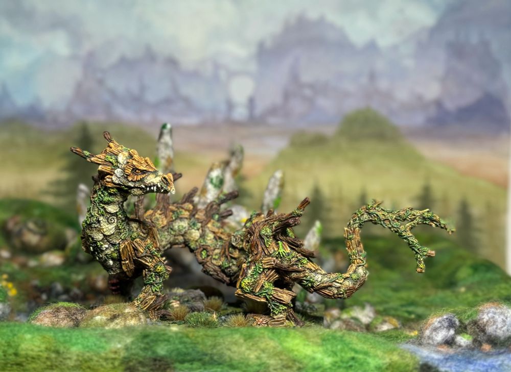 A painted forest dragon miniature on needle felted terrain with a painted landscape background.
