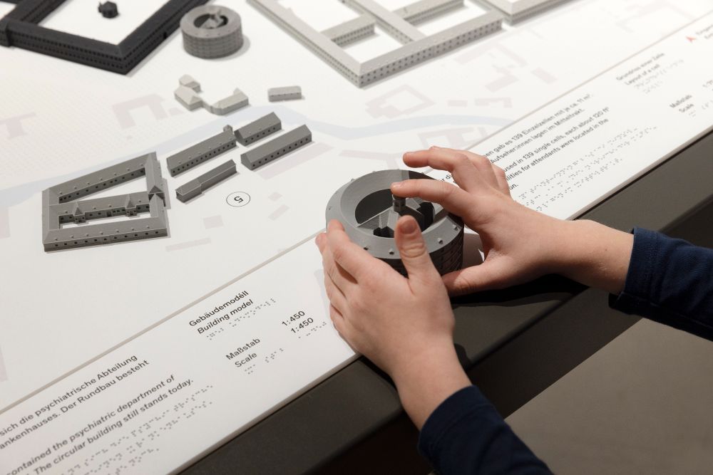 Bild zeigt einen taktilen Plan mit Gebäuden, der barrierefrei für blinde Menschen benutzbar ist - Erklärungen sind auch in Braille-Schrift verfügbar