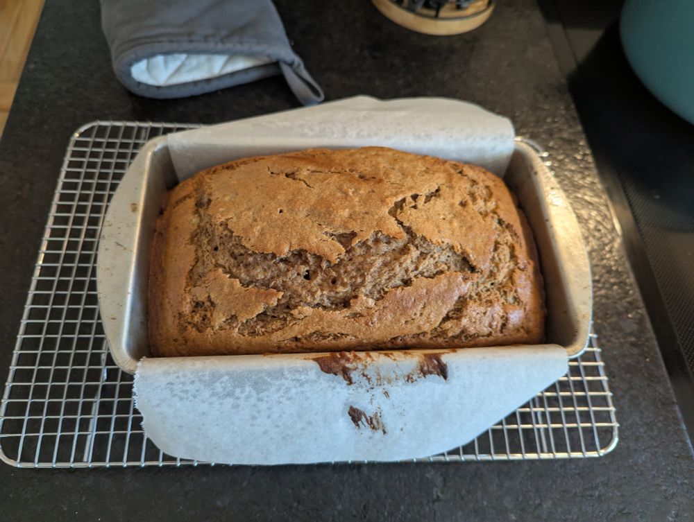 Freshly baked loaf of banana bread, still in the aluminum pan. The pan is sitting on a wire cooling rack on a black, stone countertop 