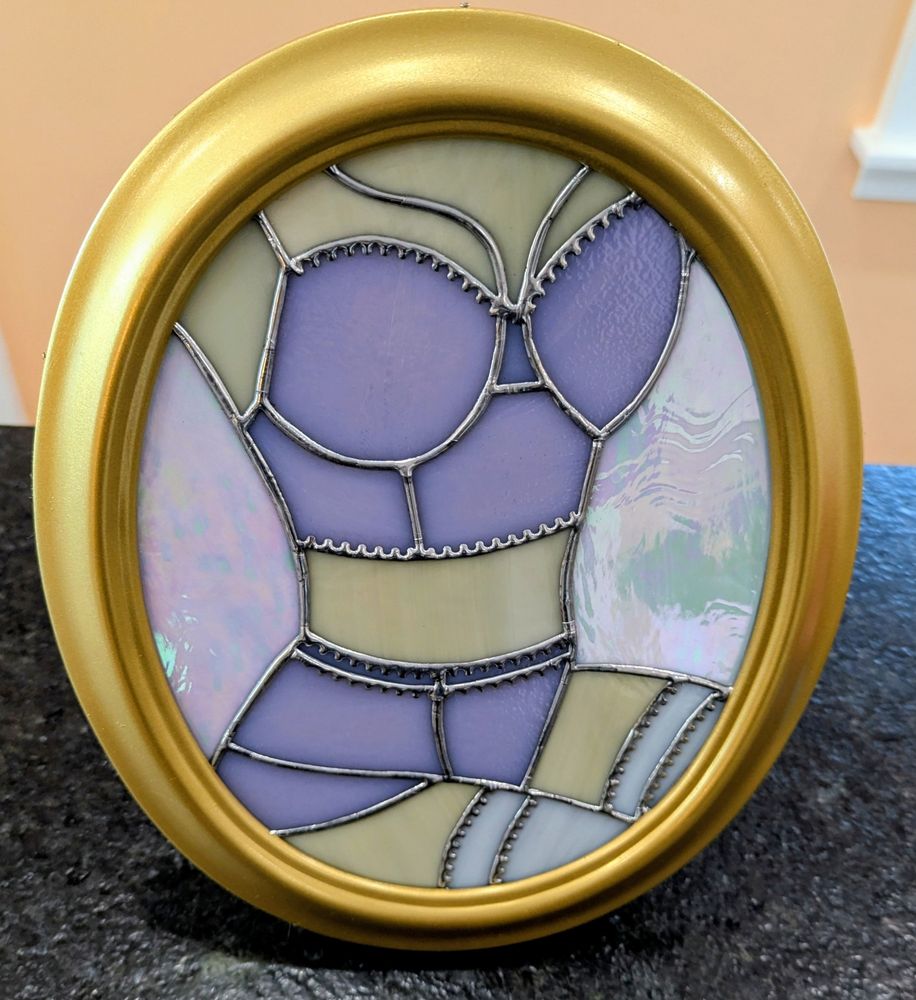 Stained glass piece depicting a lady's torso. She is seated with her legs off to the right side of the frame. She is wearing pink lingerie with white stockings. The piece is in a gold, oval frame