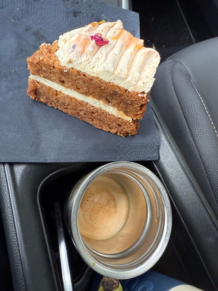 Stukje worteltjestaart op een servetje en een beker met havermelkcappuccino. Op t middenconsole in de auto