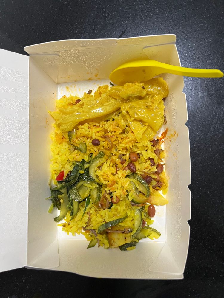Indonesian yellow rice with vegetables and peanuts in a white box with a yellow spoon in it on a black table 