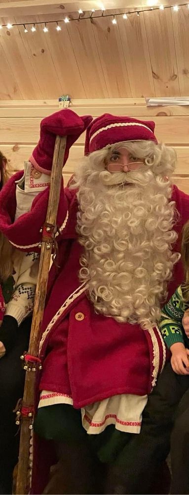 Imposter Santa. A young 20 something male with black eyebrows and an obviously fake beard. He is wearing an ill fitting red coat with a white undershirt, black pants and has a wooden walking pole in his red gloved hand. 