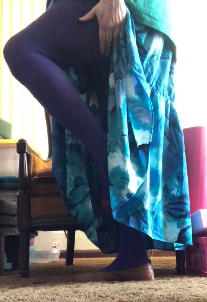 Woman in an ankle length flowing skirt that is watercolor themed in splashes of greens, blues, and whites. She also is wearing purple tights and beige flats. One leg is raised and the skirt is pushed aside exposing most of her calves and thighs.