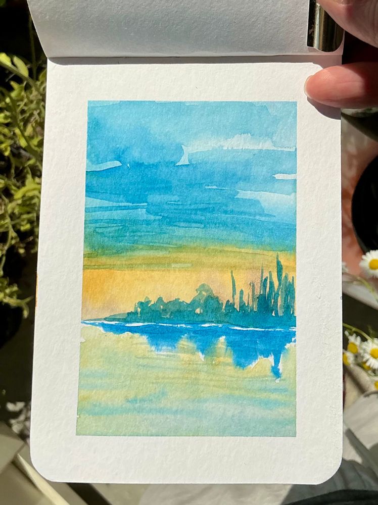 a small island at dawn with a blue sky fading into yellow on the horizon that's covered with trees and bushes, the water reflects in the water in dark blue. In watercolour on A6 paper