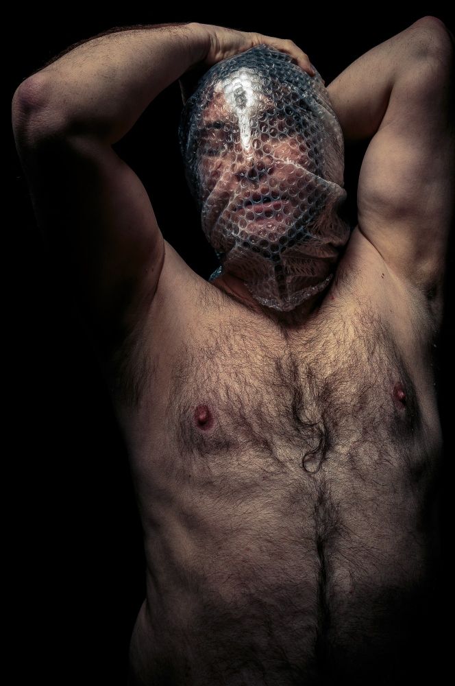 A nude man photographed from his navel up stands with his head wrapped in bubble wrap. 