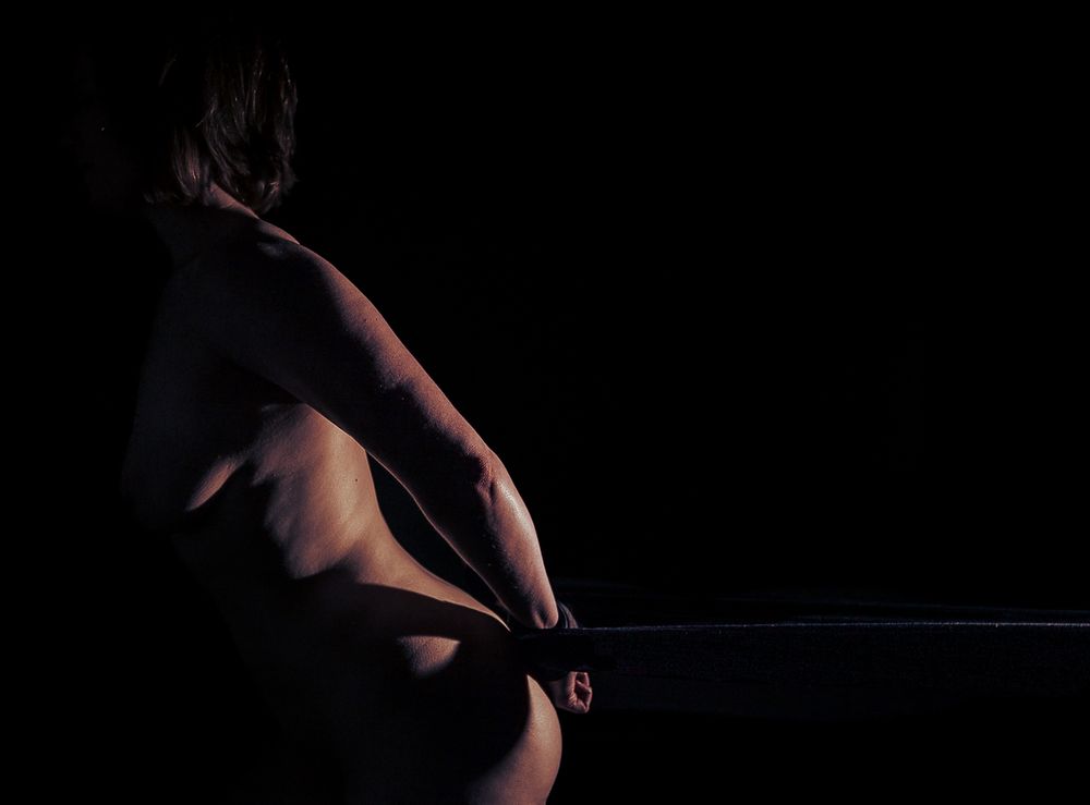 A nude woman stands in profile with her hands tied behind her back. Her body takes up half the frame. The restraint is stretched tight, taking up the other half of the frame behind her. It disappears past the edge of the photo. She is lit from the side and the front of her body falls into darkness. 