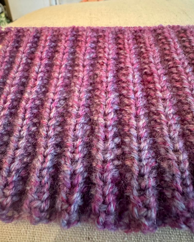 A close-up photo of the knitted rib fabric which is made in a purple hued Yarn.