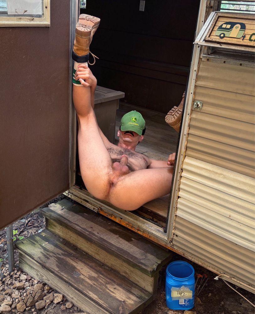 A naked man on his back exposing his hole in a trailer door, a ball cap pulled over his eyes and a tip jar on the ground