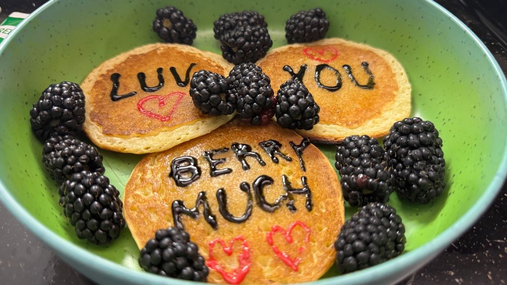 A photo of mini-pancakes with blackberries on a plate.  The pancakes have hand-written words that say “Luv you berry much” with black icing, and read heart motifs written in red icing.  This is my husband’s breakfast I make him most workday mornings.  Foodmaking is a love language for me.