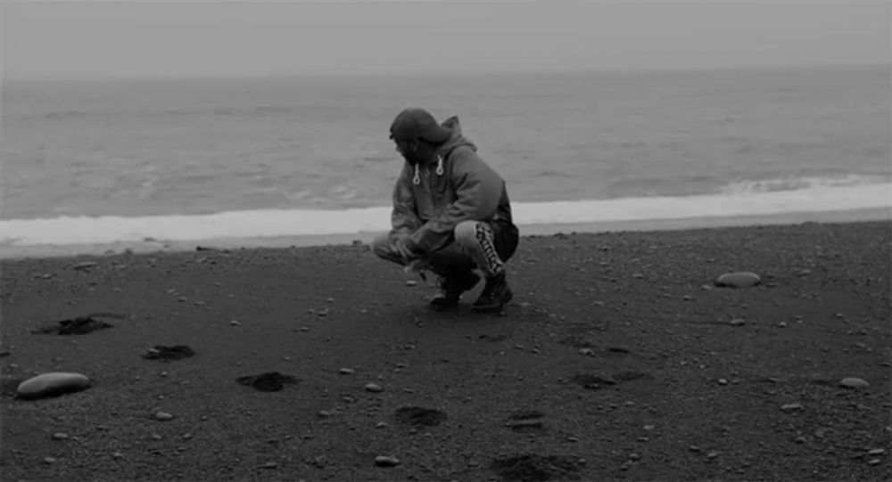 a black and white photo of a man squatting on the beach near the ocean