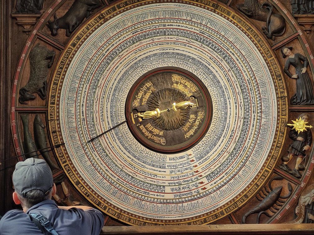 Mensch mit blauem Hemd und blauer Schildmütze lehnt links unten im Bild mit Armen auf Brüstung und betrachtet kreisrundes Kalendarium der Astronomischen Uhr in der St. Marienkirche zu Rostock aus dem Jahr 1472, das formatfüllend in der Bildmitte abgebildet und in weiß-braun-gold-Tönen gehalten ist.