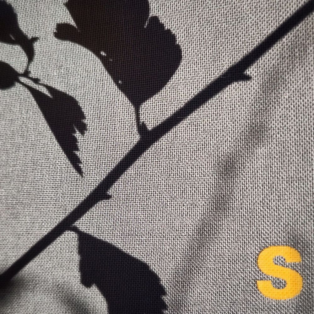 Scharfe und unscharfe Schatten von Blättern und Zweigen vor grauem Leinengewebe, in das ein gelbes 'S' geprägt ist