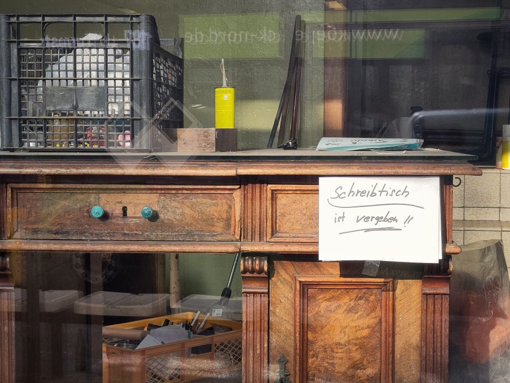 Ansicht eines alten Schreibtischs (Vintage) in einem Schaufenster. Handgeschriebenes Schild "Schreibtisch ist vergeben!!". Verschiedene weitere Gegenstände und Plastikkiste, alles deutet auf Geschäftsaufgabe hin.