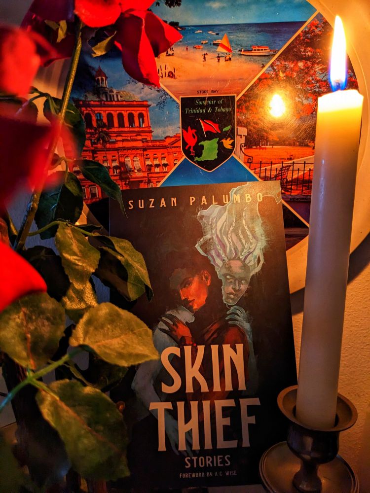 Suzan Palumbo's book Skin Thief surrounded by a Trinidad souvenir plate, a lit candle and roses. On the cover of the book is a brown person being embraced by a ghost.