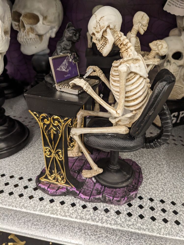 Figurine of skeleton sitting infront of a desk working on a laptop. A black cat looks on from behind the computer.