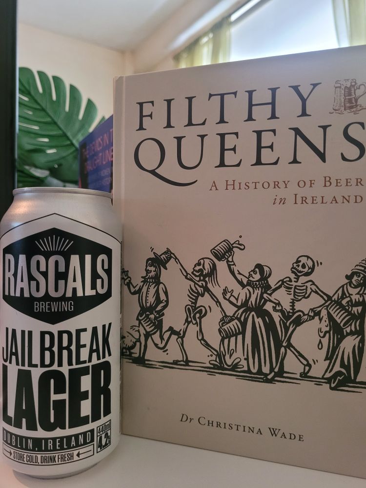 Photo of the book 'Filthy Queens: A History of Beer in Ireland' by Dr Christina Wade. The cover features a beige background and a dans macabre. It is standing up next to a silver van of Rascal's Jailbreak Lager with a plant and book in the background