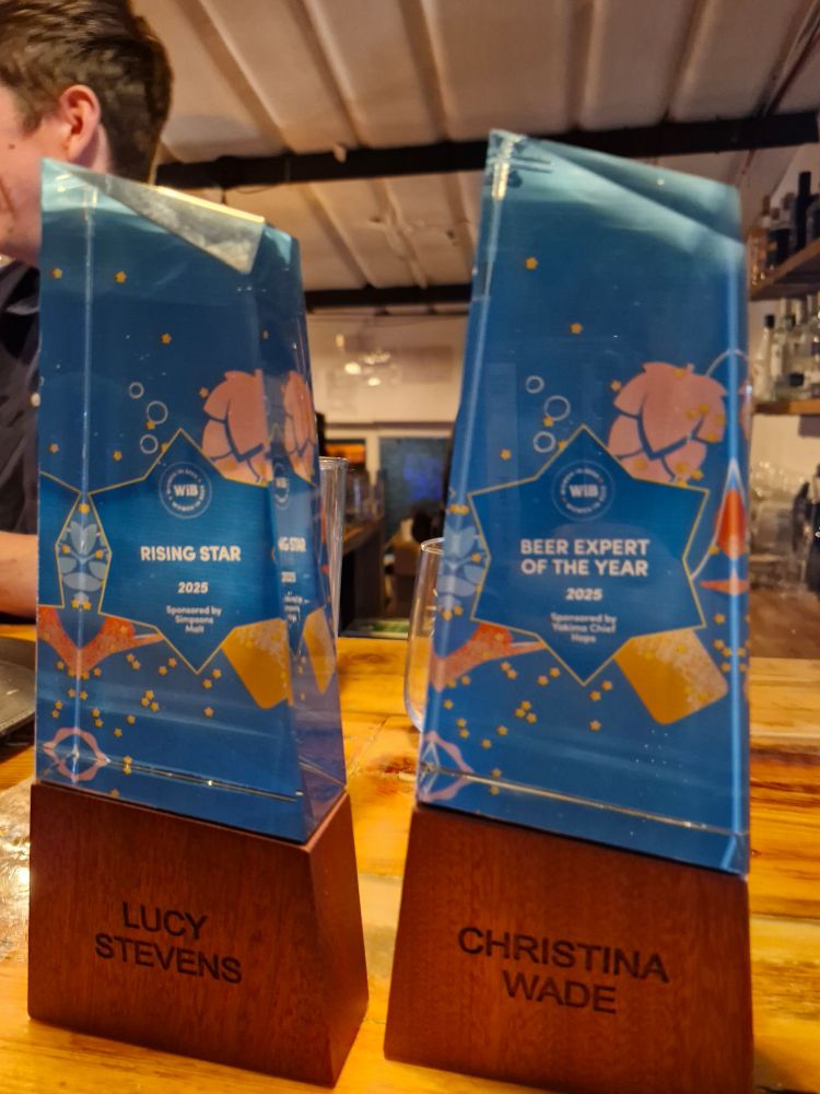 Two awards sit next to each other on a wooden table. One is for Lucy Stevens of Closet Brewing who won Rising Star. The other is for Christina Wade who won Beer Expert of the Year