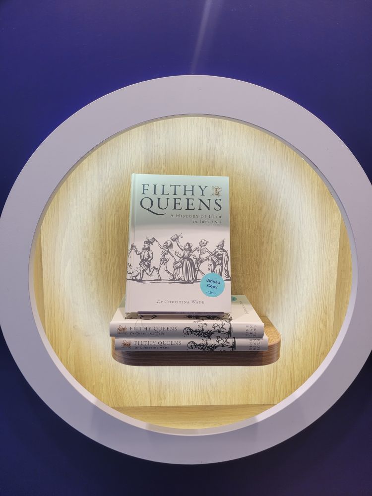 Photo of three book Filthy Queens: A History of Beer in Ireland, two stacked on top of each other and one leaning upright with the cover facing us. Books are in a circular wooden alcove with a birch background, a white circle encloses it and a blue wall.