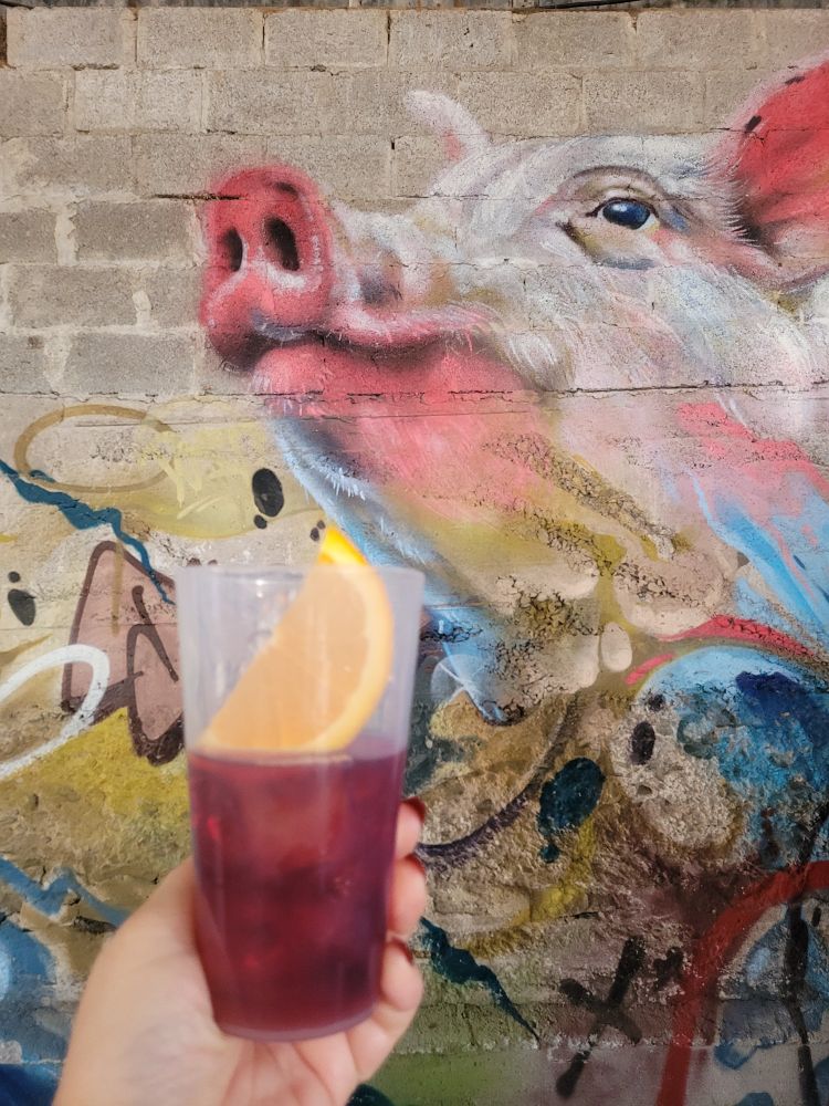 Photograph of wall with pig painting. A woman's hand holds up glass of red Mead with an orange slice in front.