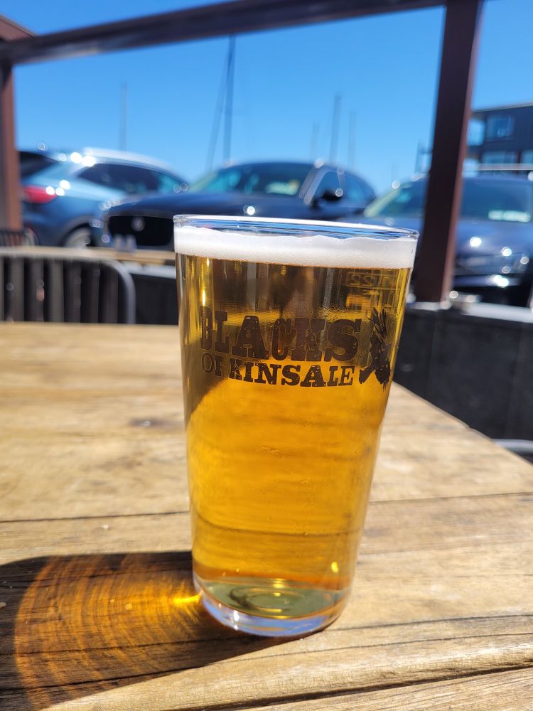 Photo of a pint of Blacks of Kinsale Lager on a wooden table, outside one very sunny day. In the background are cars and sailing masts