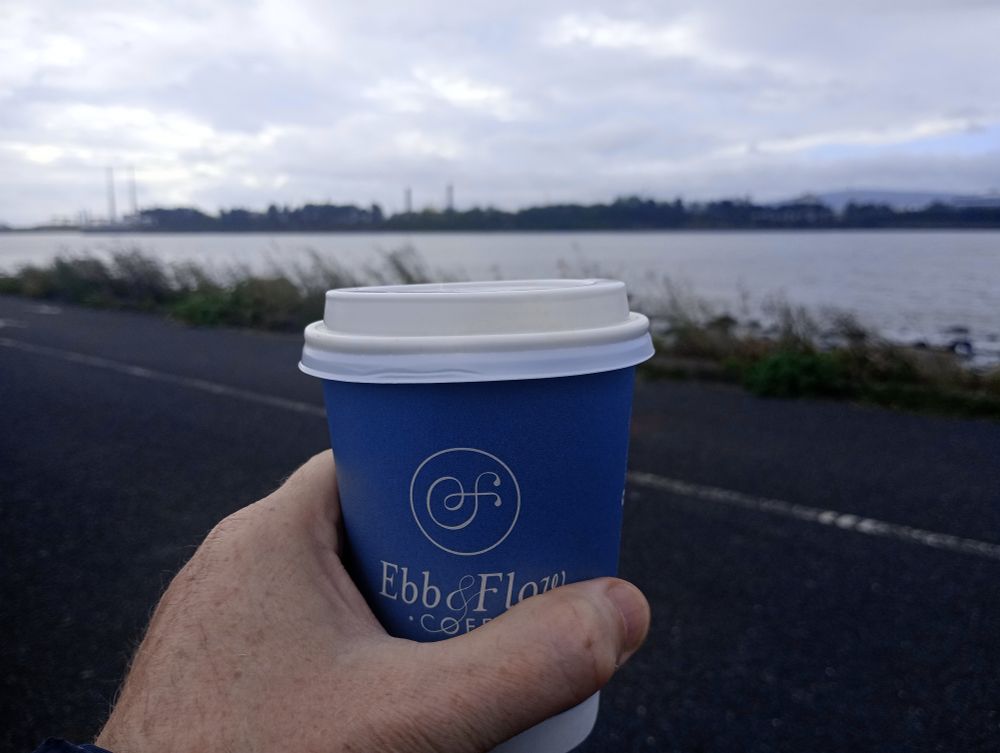 Photo of hand holding a takeaway coffee with Clontarf bay behind.