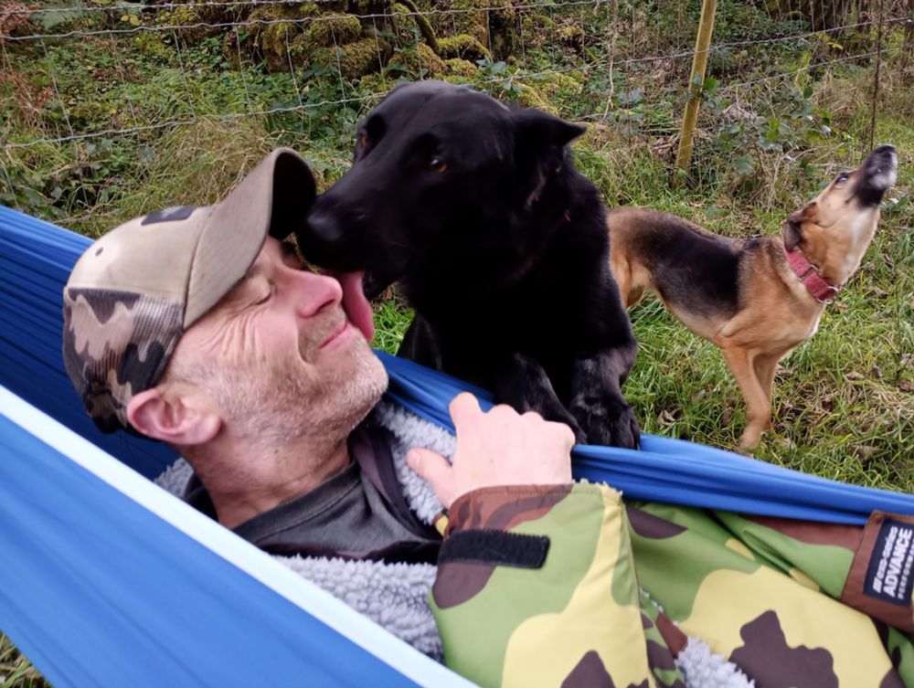 Photo of Obie, a black German shepherd, licking Leon's face as he lies in his hammock with Jessie barking in excitement 