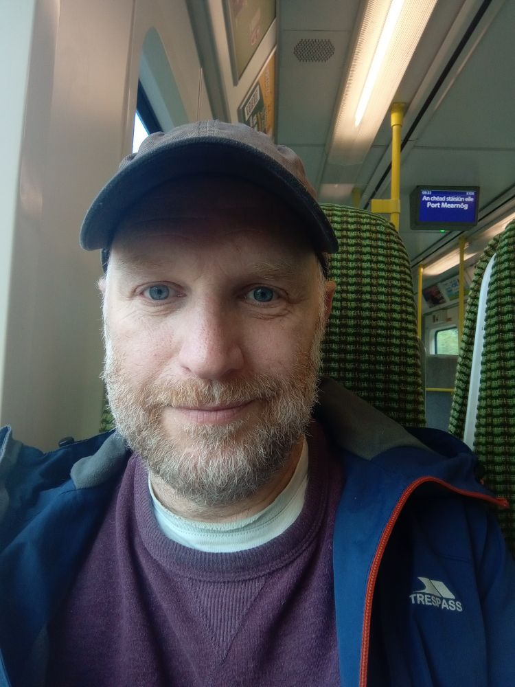 Selfie of Bob on the Dart with half a smile & a blue cap & a short ginger beard that's half grey. 