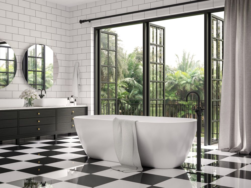A luxury bathroom featuring a black-and-white tile floor, white tile walls, black cabinets, dual sinks, and a soaking tub with 3 large windows open to the lush outdoor landscape.