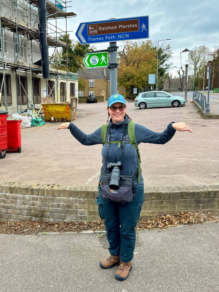 A woman (me) with a camera round her neck, by a Rainham Marshes sign, pretending to be a bird. 