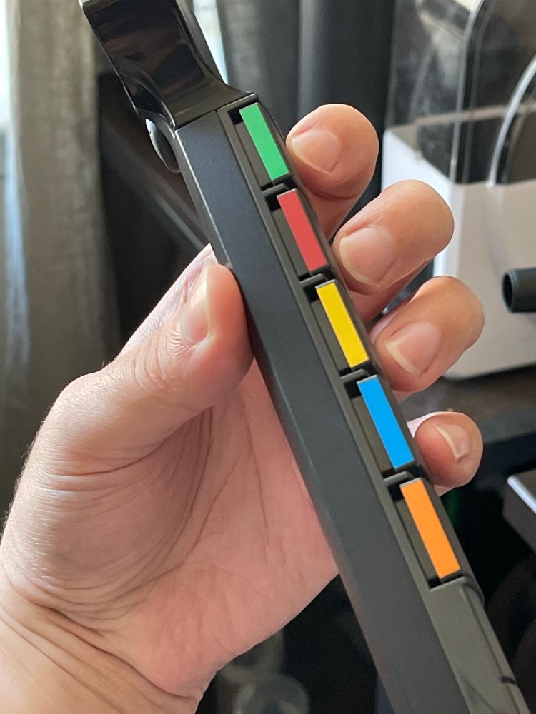 holding the neck of a guitar hero controller with the thumb tip touching the back of the neck, facing toward the fingertips. proper form is to lay the thumb on the neck with the tip facing toward the top of the guitar, allowing it to lay in the palm