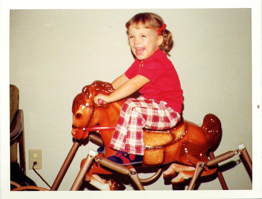 Paula as a young child with ponytails and red barettes, wearing plaid polyester pants and a red top, riding a Vintage Wonder Horse (Spring Horse).