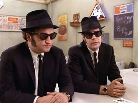 Filmausschnitt mit den Blues Brothers (schwarze Anzüge, Hüte, Sonnenbrillen, an einem Tisch) 