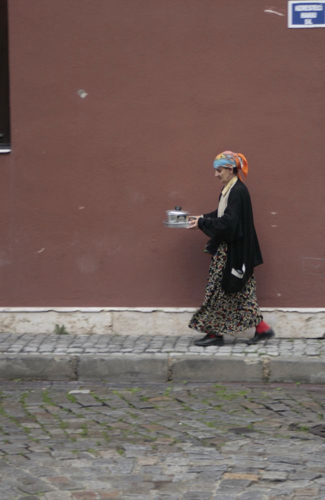 Frau mit Kopftuch läuft auf einem Fußweg, sie trägt ein Tablett mit einem Topf oder Samowar o.ä.