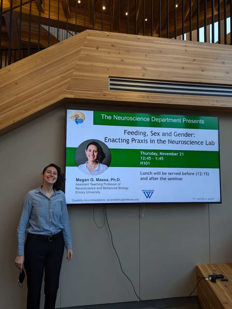 Megan, a white person with a half shaved head and a half head of black curly shoulder length hair, wearing a blue long sleeve collared and button down blue shirt and long black pants, stands smiling next to a digital poster reading "The Neuroscience Department Presents: Feeding, Sex, and Gender: Enacting Praxis in the Neuroscience Lab" along with event details.