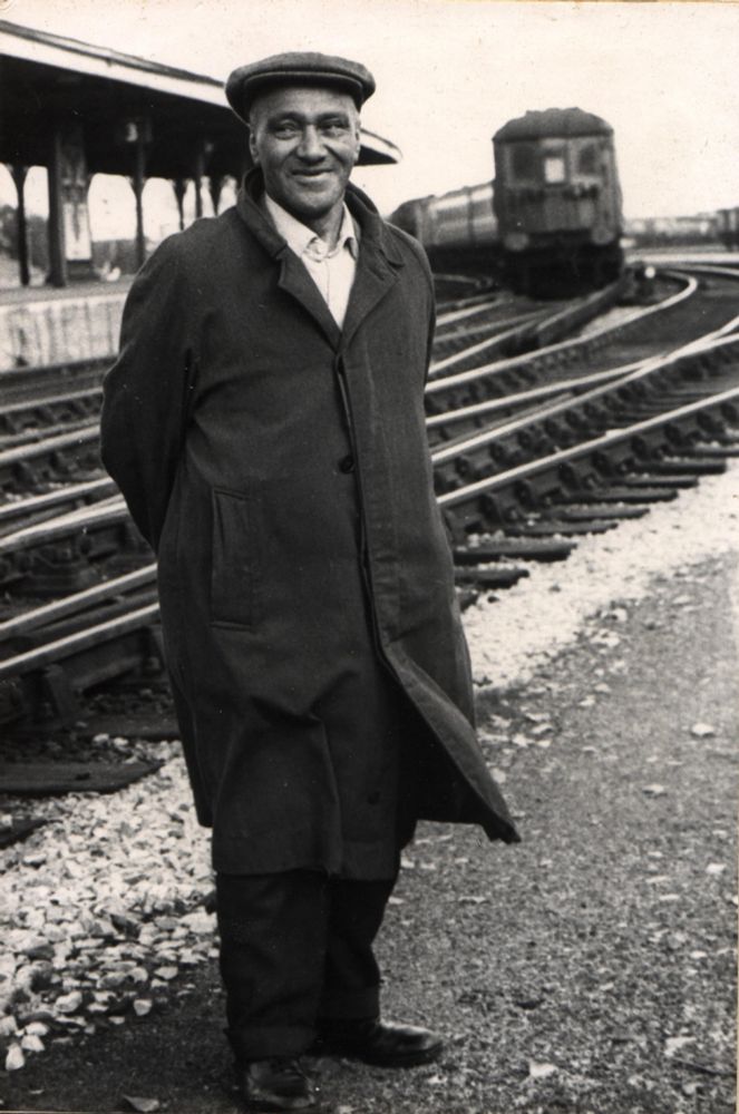 Man in coat and flat cap stands smiling to someone off-camera; an electric train and platform is behind him. Courtesy Chris Cornell.