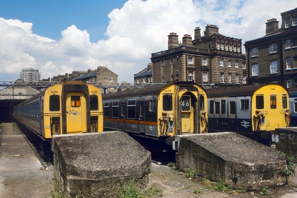 Colour photograph of British Rail era electric slam door multiple unit stock in sidings in an urban environment, under a blue cloudy sky.