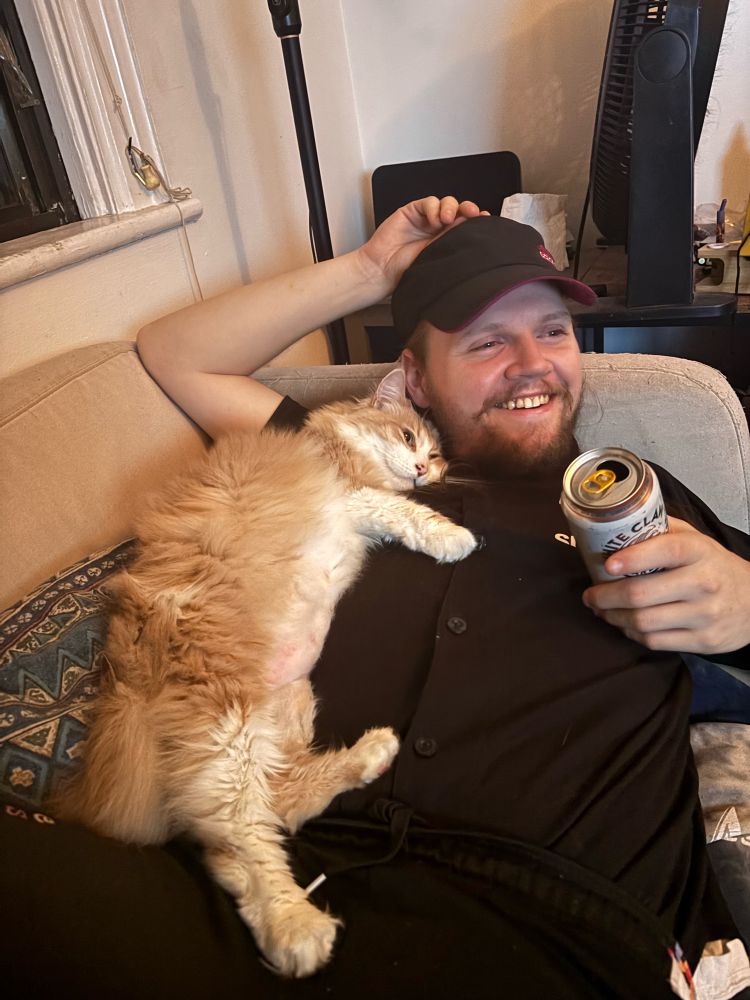 a ginger cat hugging a person on a couch who is smiling and holding a drink.