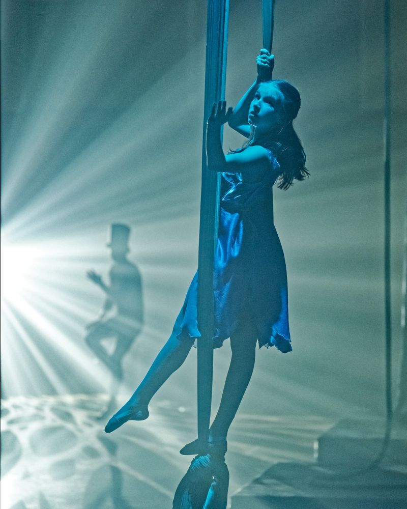 A young girl is standing in a fabric suspended from the ceiling. Blue light dominates with a bright spotlight in the rear silhouetting a man dancing and wearing a tall top hat.