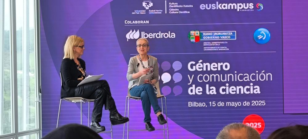 Las dos ponentes sentadas en una sillas altísimasv frente al pantallazo morado del encuentro.