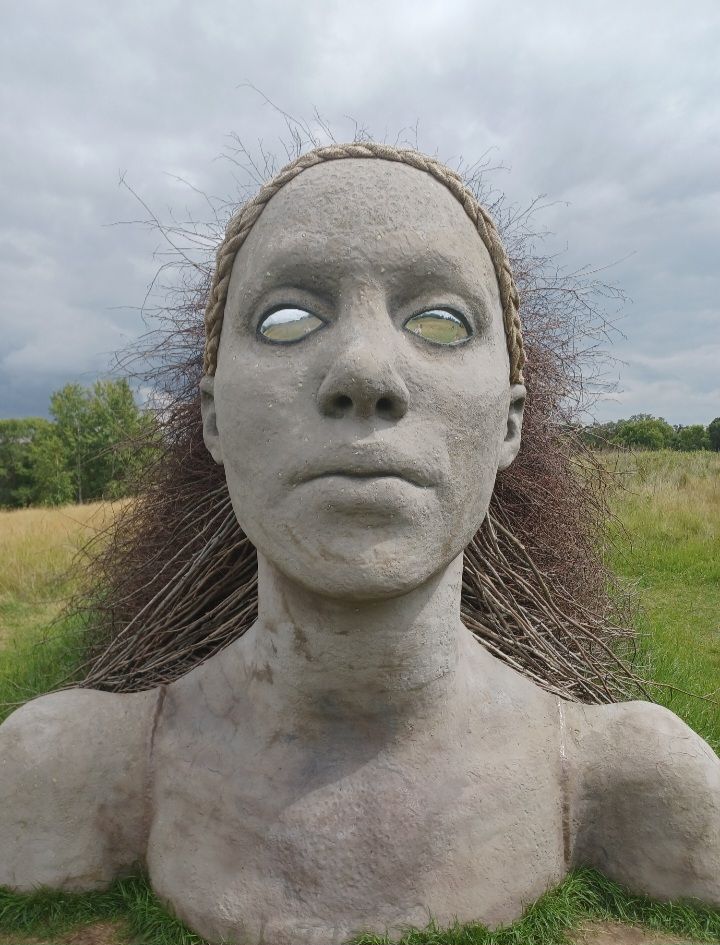 An outdoor sculpture of a woman from the shoulders up, she rises from the prairie with her hair, made up of twigs and branches, flowing out behind her. Her eyes are mirrors. 