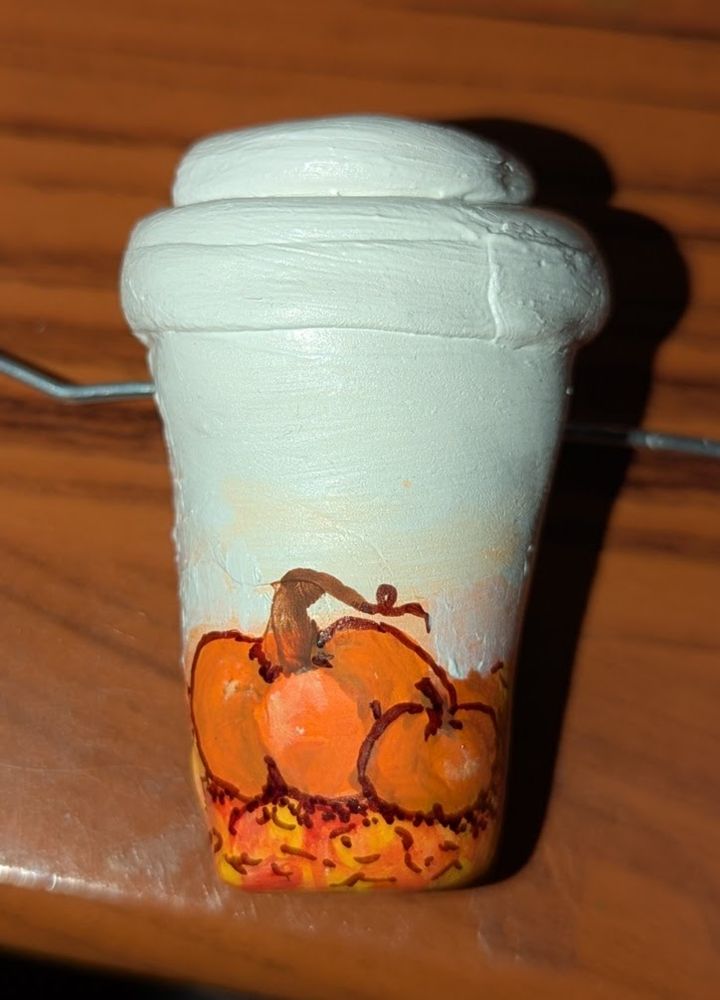 A small clay take away coffee cup painted with a pair of pumpkins on a field of fall leaves.