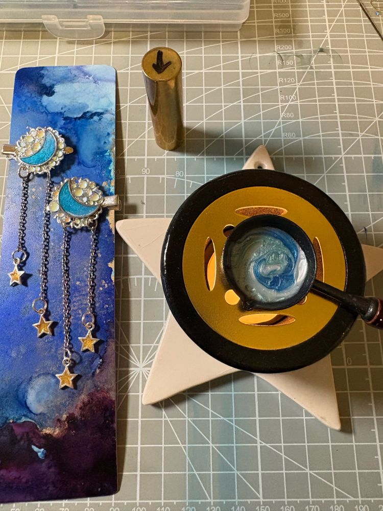 A blue sky sheet with two hair clips attached sitting next to a wax burner.  The hair clips have moons and iridescent beads and silver chair with yellow stars hanging from it.