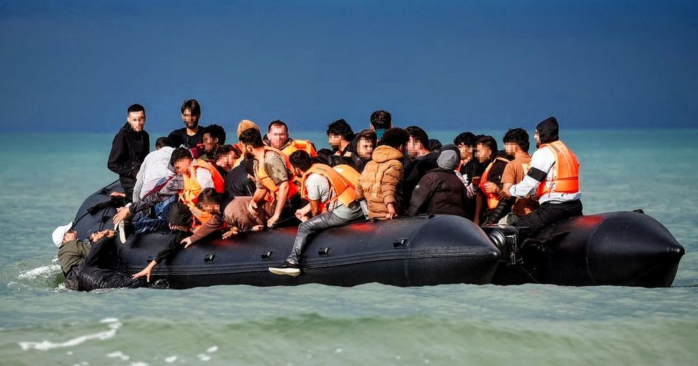 Black rubber boat on open water dangerously overloaded with passengers, only a few of whom have life jackets. Some are leaning out, reaching for a person in the water.