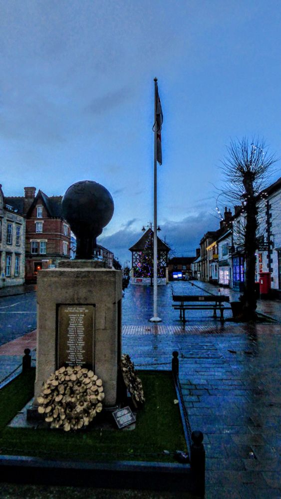 Wootton Bassett War Memorial, a limestone plinth topped by a bronze world globe held aloft with a narrow pedestal of flat hands. Two bronze poppy wreaths lean against the plinth which bears the names of the fallen in white on bronze. Behind is a limp flag on a flagpole snd behind that, in thr distance, is the old town hall.
