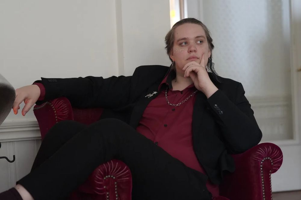 A person lounging in a red chair, they are wearing a black suit with a red shirt