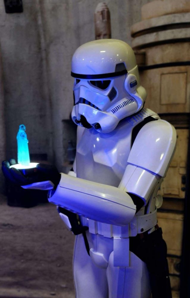 An a new hope Stormtrooper looking at a holovideo of Darth Vader that he's holding in his left hand. The background is a dirty Tatooine-style setting. 